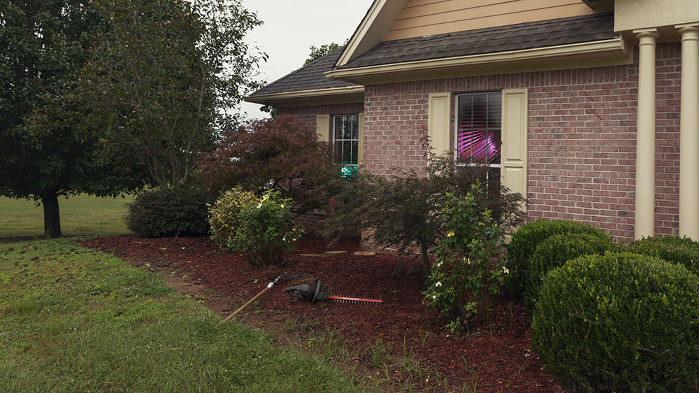 garden tools in mulch bed by house, windows show glowing inside house