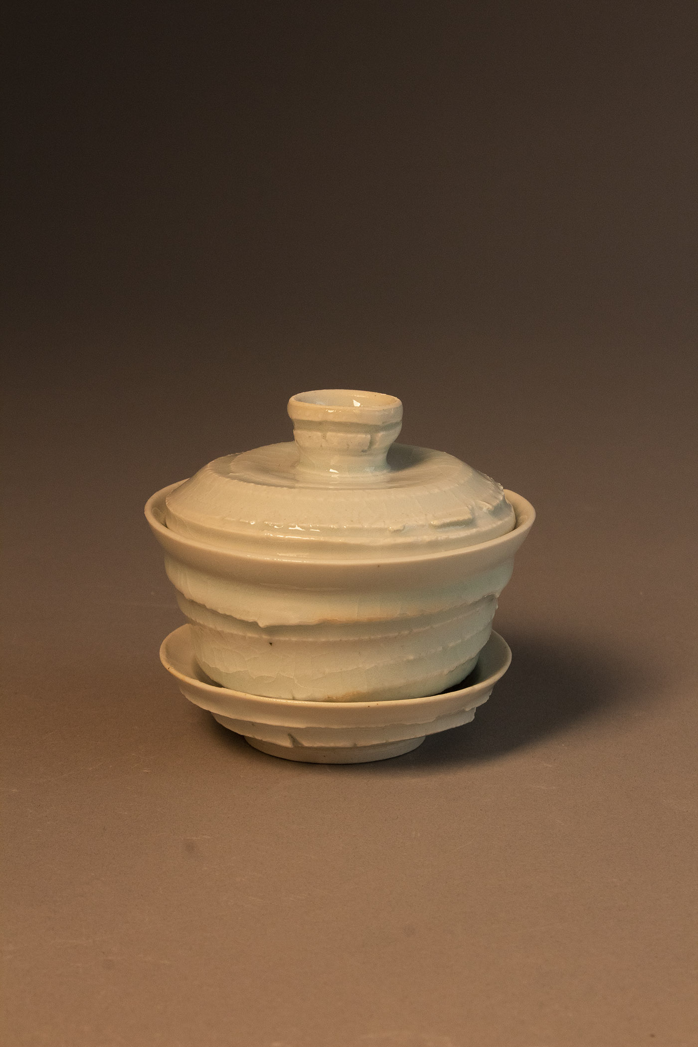 A three-part vessel for steeping tea – consisting of a saucer, a bowl-shaped cup with an outward-flaring lip, and a lid with a knob. The exterior surfaces have some slightly abrasive ridges and texture from the stretching of the clay during throwing. The slight roughness contrasts the delicacy of the vessel (evident in the apparent thinness of the rims of the cup and saucer), and the glassy glaze.