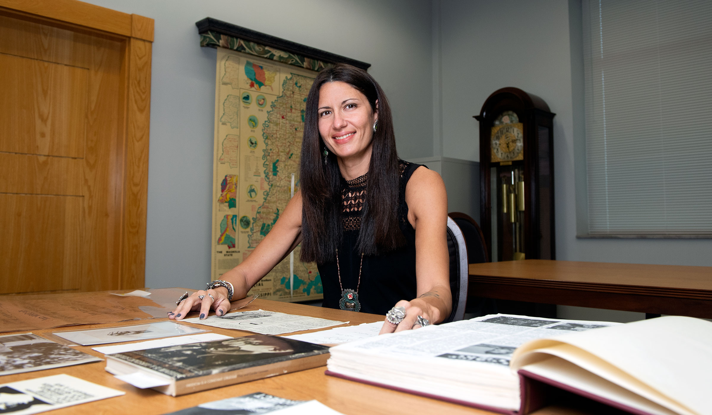 Dr. Altomonte sitting at table with books and papers smiling at the camera while wearing a black tank top.