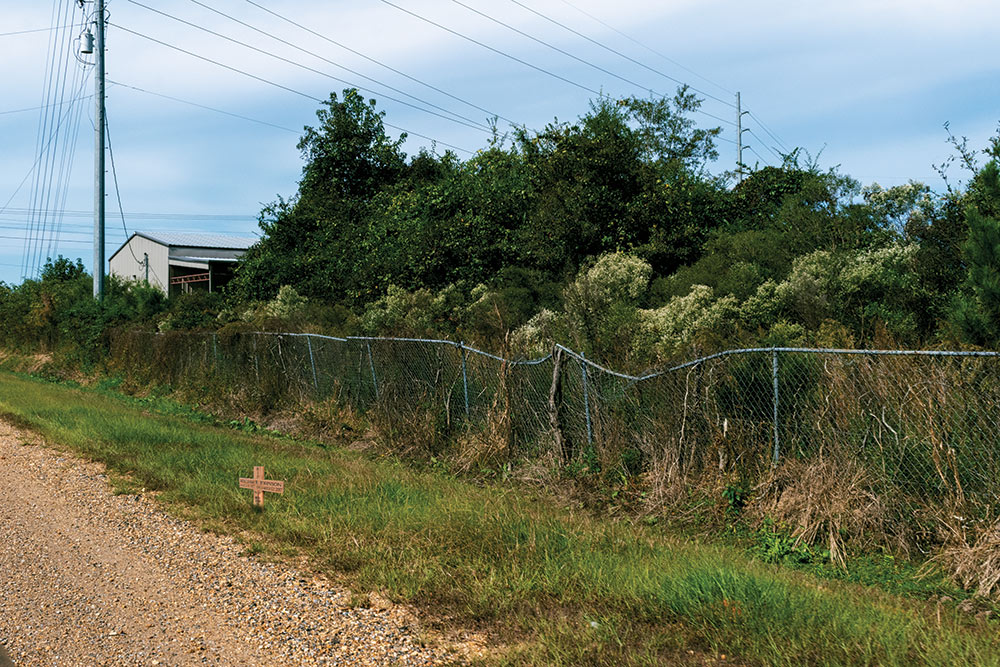 cross on side of gravel road. chain link fence that's bent is seen behind it