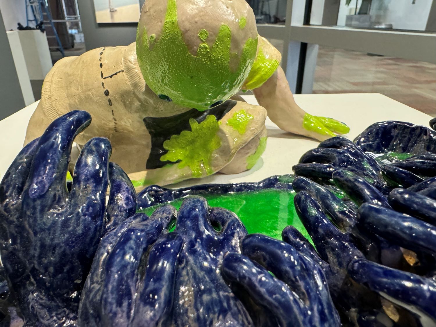 Sculpture of a human figure with a dripping green face, with hands positioned over a pool with vibrant blue texture elements and a shimmering green surface.