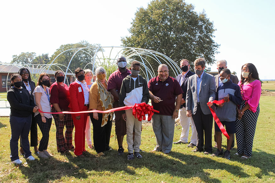 People stand in front of new community garden cutting ribbon: from left, Esperanza Colon, student; Barbara Lucas, principal; Emiline Brown, science teacher; Jamesha Keithley, science teacher; Susie Williams, district curriculum coordinator; Allison Poindexter, Delta Health Alliance; Suzanne Powney, MSU associate professor of art; Alexis Hamilton, MSU Extension agent I; Caleb Davis, student; Rev. Jesse King, district superintendent; Scott Willard, interim dean of MSU’s College of Agriculture and Life Sciences; Cory Gallo, landscape architecture professor; Sadik Artunç, professor and head of MSU’s landscape architecture department; Marquis  Wafford, student; and Rickeya  Brown, science teacher. 