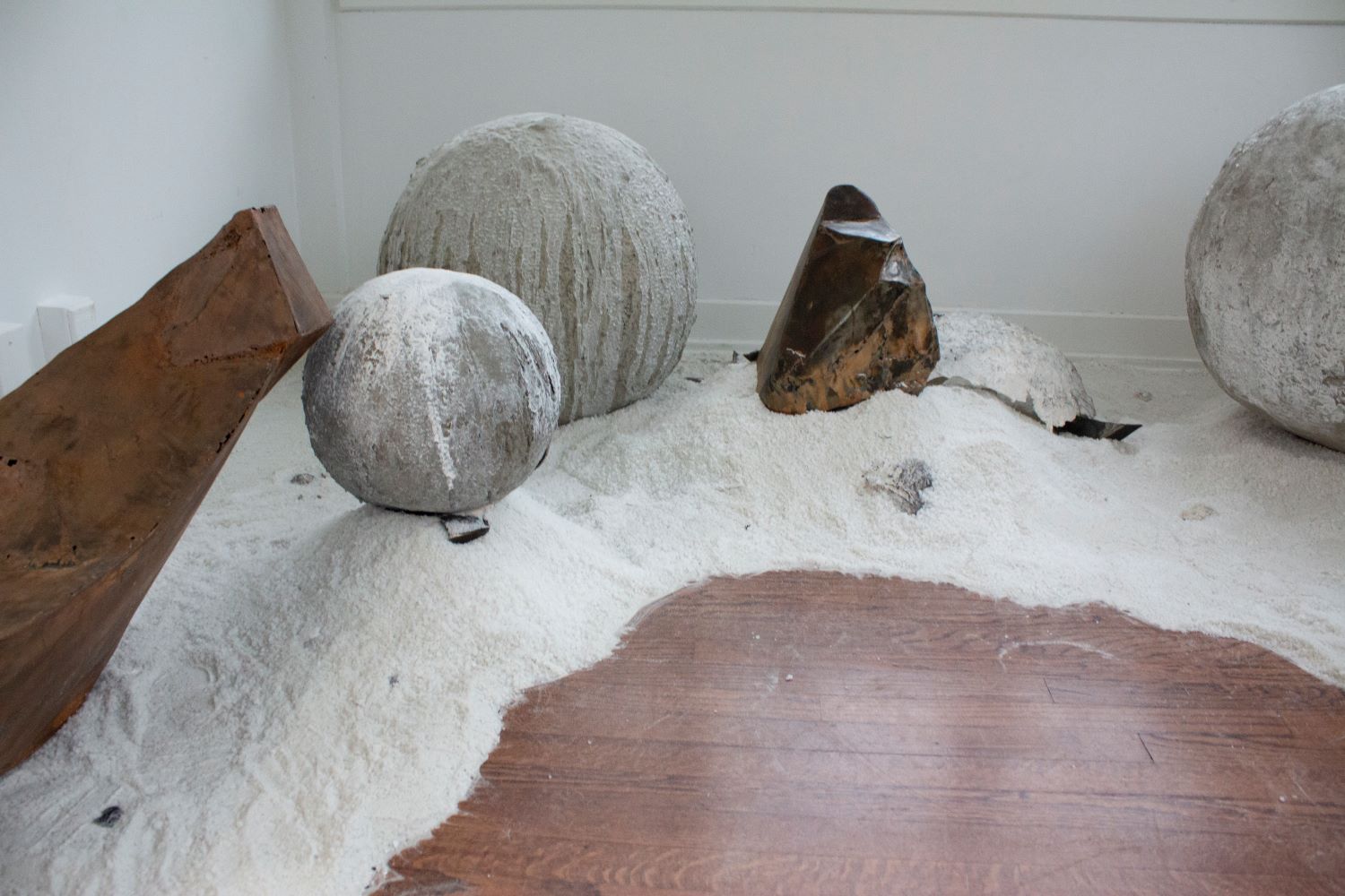 3 Large concrete orbs and 2 triangular rusty metal sculptures sitting in sand at multiple elevations. 