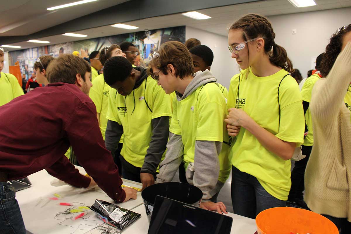 male BCS student explains their tape measure challenge to a large group of the students who attend the career fair (in yellow shirts) 