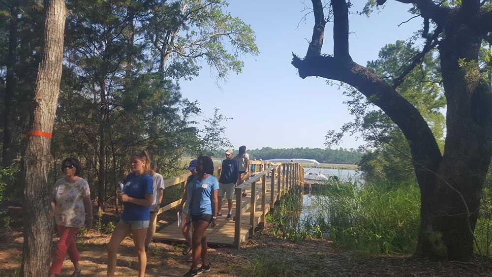 Internship boat trip and orientation. A group walks over a bridge from the water and boat into a wooded area.