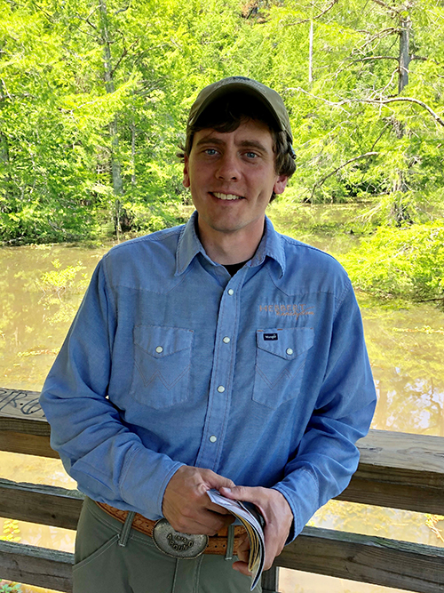 Jake Hebbert (Photo submitted) stands wearing a blue shirt and tan baseball hat in front of a lake with trees. He is holding a booklet.