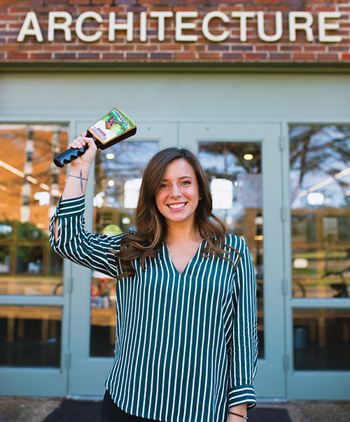 McKenzie Johnson stands under Giles Hall's "architecture" signage and rings cowbell with her right arm