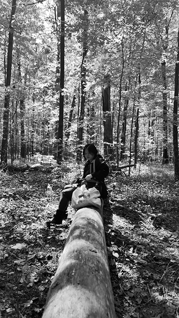 Black and white photograph of a figure sitting on a fallen tree in the woods