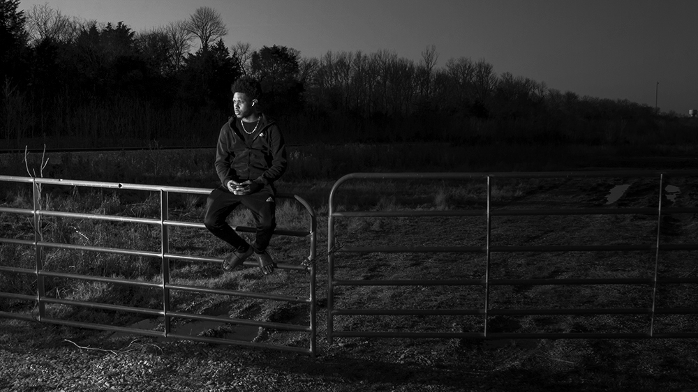 A photograph of a boy sitting on the fence of a gate. 