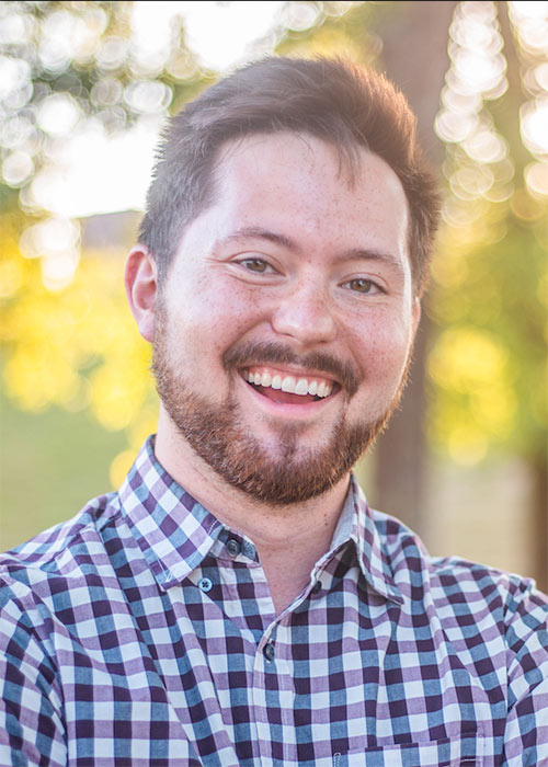 Asher Paxton headshot - outside with greenery in background wearing blue checkered dress shirt