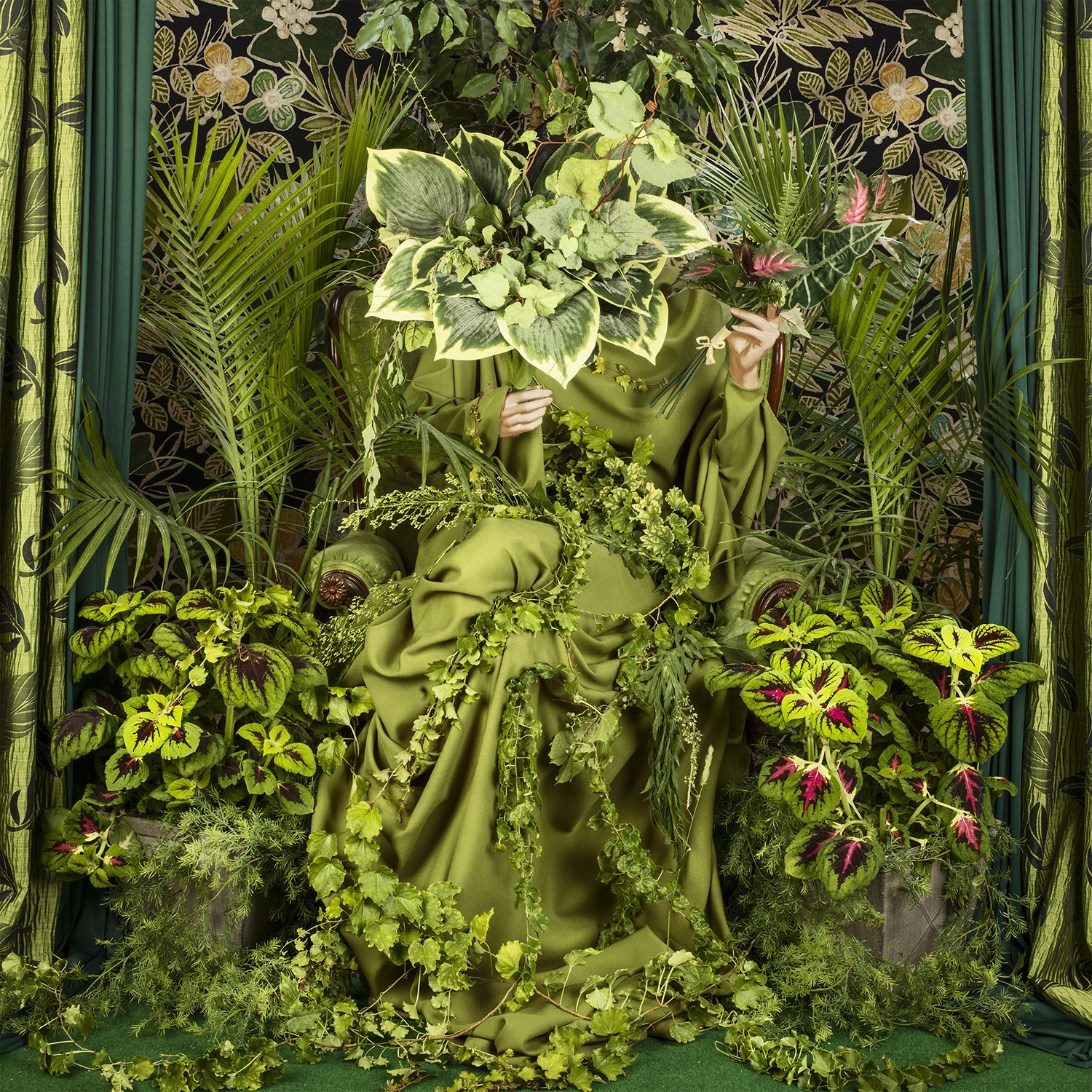 Photograph of a seated figure covered in green leaves and plants.