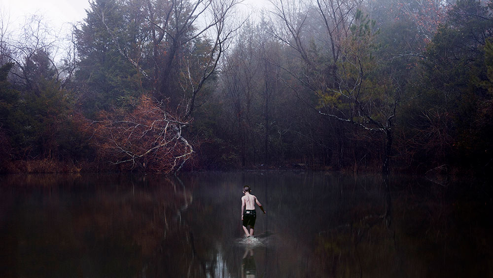 boy wearing dark shorts and no shirt stands in body of water surrounded by trees