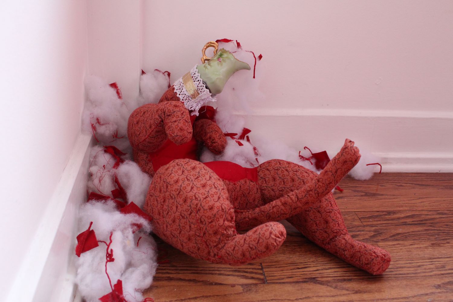 A red fabric cat sculpture with a vintage cream dish for a head pours polyfill out of its head. It sits in its own stuffing