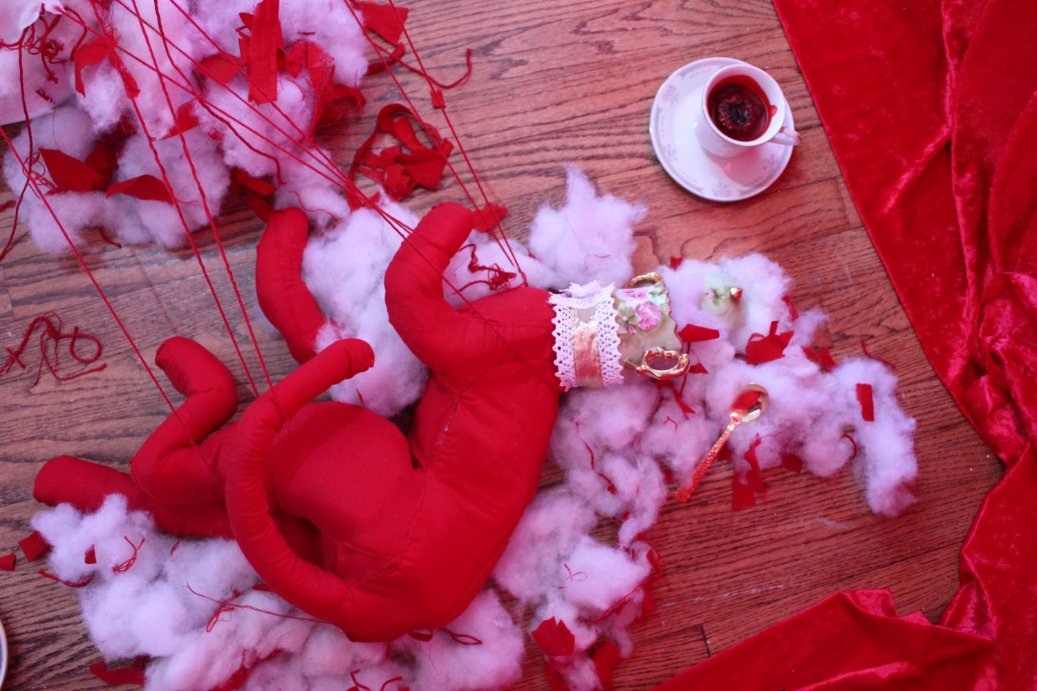 A red fabric cat sculpture with a vintage sugar dish for a head spills polyfill out of its head. It sits in its own stuffing. It is tied in red threads, pulling or playing