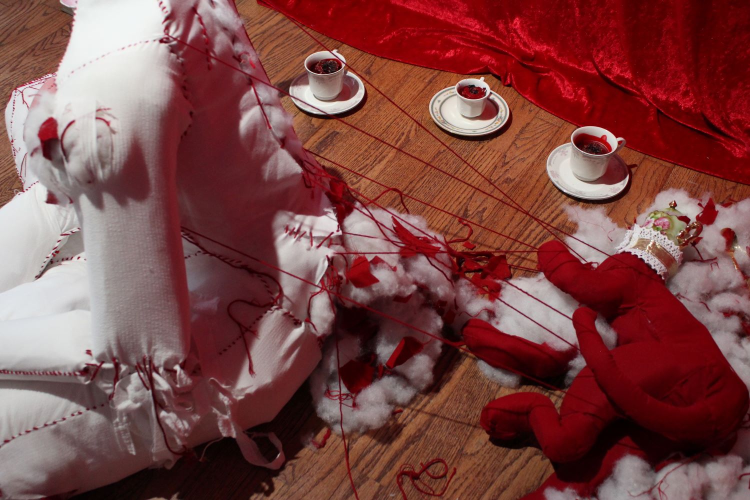 A red fabric cat sculpture pulls strings at the back of the figure, unfurling the stitches of the spine. The base of the spine has become undone, pouring stuffing out.