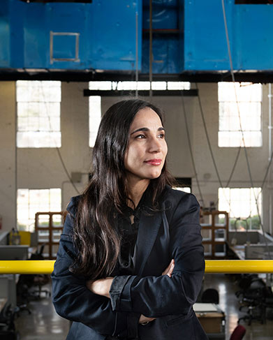 Silvina Lopez Barrera stands with arms crossed leaning against yellow railing on second floor of Giles Hall at MSU overlooking first-year studio space