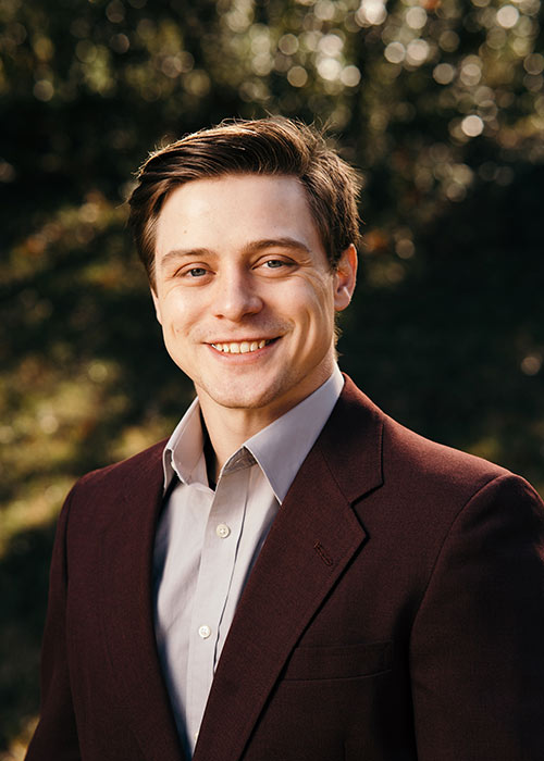 Steve Dragoo headshot - wearing maroon suit - outside with greenery in background