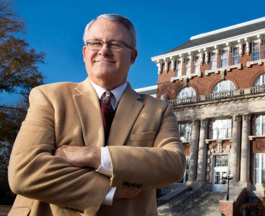 Tim Muzzi poses in front of brick campus building, arms crossed