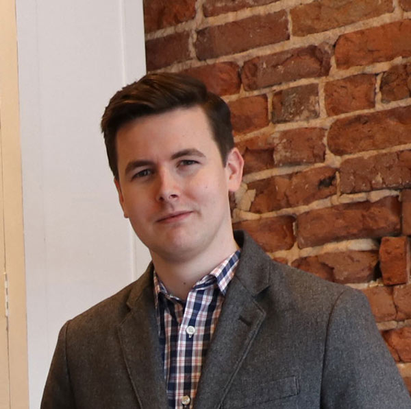 Nathan Thomas smiling at the camera, wearing a plaid shirt and grey blazer.