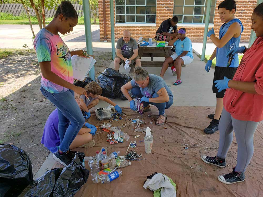 Intern Tasia Williams records data on litter collected at various sites in Magnolia Bayou Watershed. Students are sorting various trash pieces on a tarp while Tasia stands with paper and pen