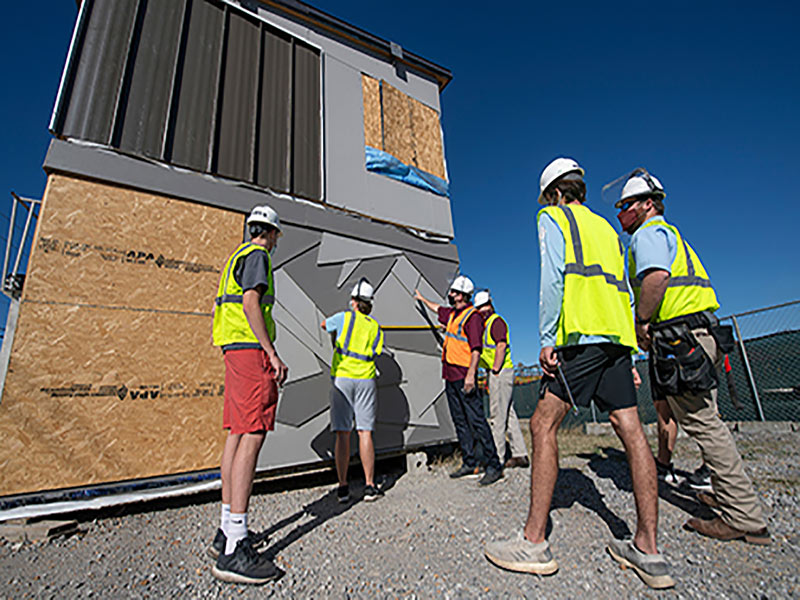 Six men in construction work gear measure and look on at a tiny, grey house they are working on.